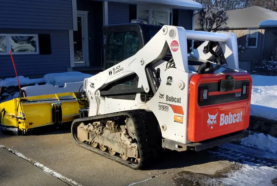Rear and side view of a Bobcat T650 compact track loader beside snow, with a snow attachment.