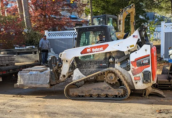 Side view of a Bobcat T66 R-Series carrying paving blocks on pallet forks at a construction site.
